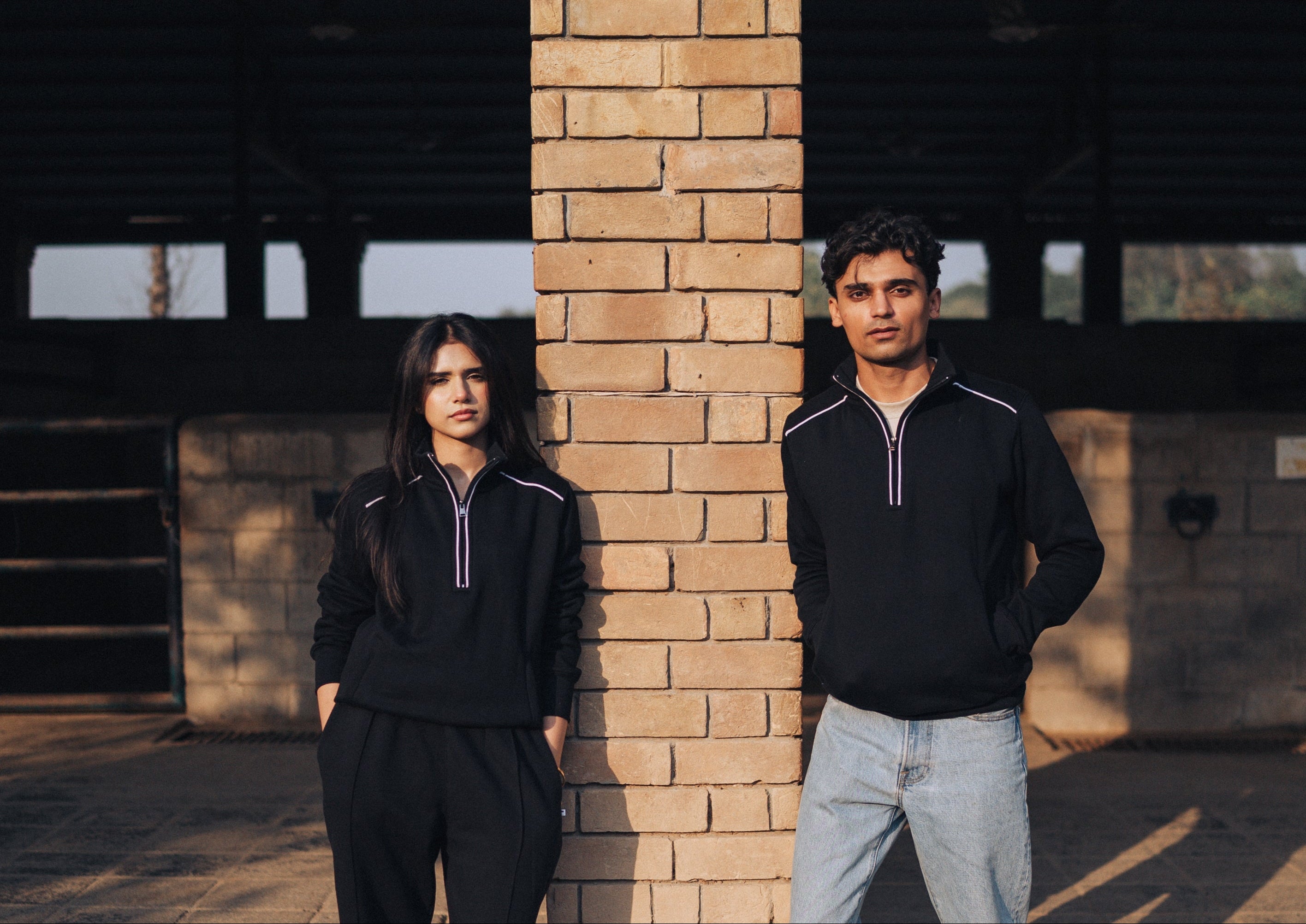 Two people wearing matching black outfits standing against a brick wall.